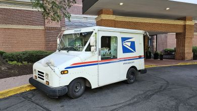 A U.S. Postal Service van is parked under an apartment building canopy entrance.