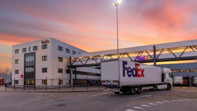 A FedEx tractor trailer truck drives into a FedEx terminal with a pink sky at dawn in the background.