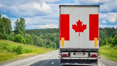 Rear view of a white semi-trailer truck with a large Canadian flag painted on its doors driving on a rural highway through green hills and trees.