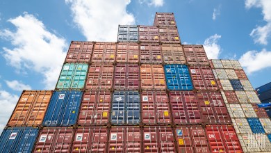 Large stack of colorful intermodal shipping containers piled high at a busy port terminal under a clear blue sky.