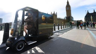 A courier steers a brown UPS electric quad delivery bike in London with London Tower in the background.