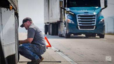 Roadside mechanic performs repair on a semi-truck trailer with warning triangle deployed and another truck in the background
