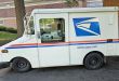 A side view of a U.S. Postal Service delivery van in front of an apartment building.