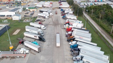 A line of trucks sitting parked in both directions at a truck stop while a single truck drives between the parked tractor-trailers.