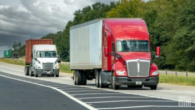 a red US Xpress sleeper cab pulling a dry van trailer with a white tractor pulling an ocean container in the background