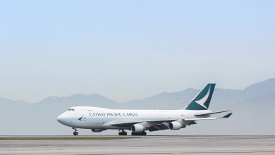 A teal-tailed Cathay Cargo jumbo jet rolls on the runway with mountains in the background.