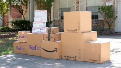 Mixed FedEx, UPS Store, USPS Priority Mail and Amazon packages stacked on a residential driveway, illustrating last-mile carrier diversification by retailers
