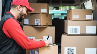 Delivery driver in a red uniform and cap reviews parcel details on a tablet at the open rear of a van stacked with cardboard boxes bearing QR codes and shipping labels