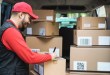 Delivery driver in a red uniform and cap reviews parcel details on a tablet at the open rear of a van stacked with cardboard boxes bearing QR codes and shipping labels