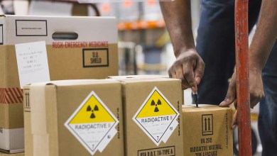 Boxes with hazmat marking for radiopharma contents are rolled on a manual conveyor into a truck.