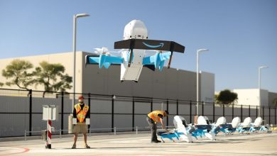 A light blue-and-while Amazon drone takes off from a line of drones with a manual handler and building in the background.