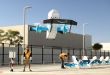 A light blue-and-while Amazon drone takes off from a line of drones with a manual handler and building in the background.