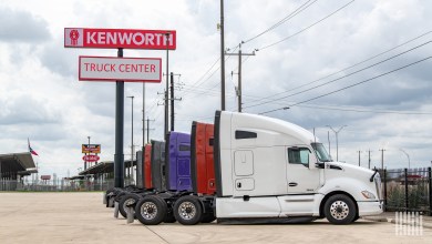 White Kenworth tractor parked at a Kenworth Truck Center dealership with a line of colorful trucks behind it and a Peterbilt sign visible in the distance