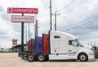 White Kenworth tractor parked at a Kenworth Truck Center dealership with a line of colorful trucks behind it and a Peterbilt sign visible in the distance
