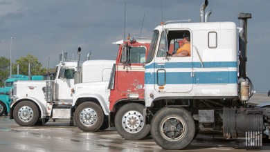 Semi-trucks parked in a lot with a driver visible in the cab of a white and blue GMC tractor