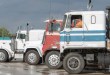 Semi-trucks parked in a lot with a driver visible in the cab of a white and blue GMC tractor