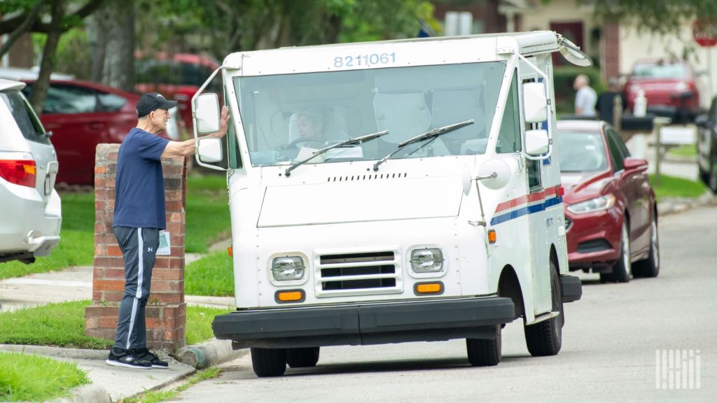 A USPS mail truck is parked on a residential street with a person standing beside it.