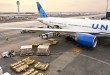 Overhead view of a United Airlines jet on the ramp at Dulles Airport on a sunny day as a tug moves cargo containers on the ground.