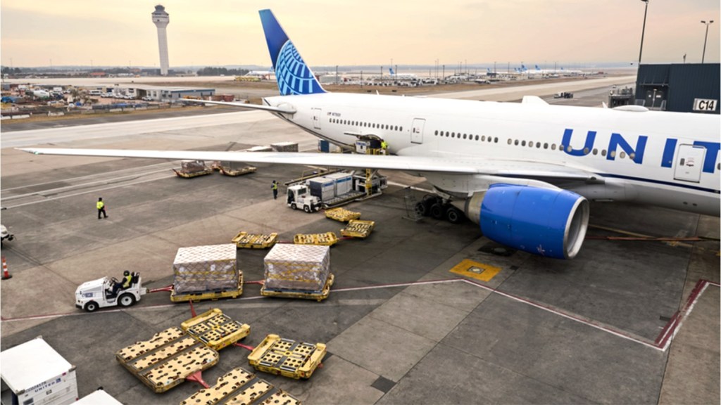Overhead view of a United Airlines jet on the ramp at Dulles Airport on a sunny day as a tug moves cargo containers on the ground.
