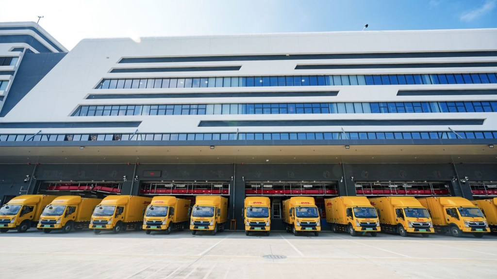 Yellow DHL trucks are seen lined up at a large air terminal as seen from the ground level.