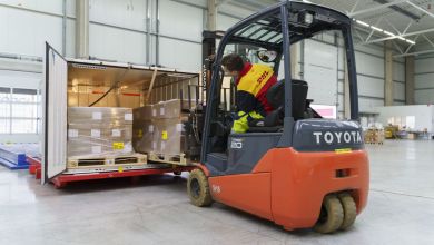 A forklift driver wearing a yellow DHL jacket loads a container with bags.