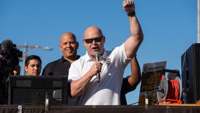 A man in sunglasses speaking into a microphone with his other fist in the air at a rally.
