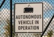 A sign reading “Autonomous Vehicle in Operation,” with a cabless truck icon and wireless symbol, zip-tied to a chain-link fence.