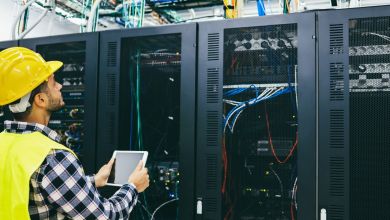 A man in a yellow hard hat and yellow vest checks cables in a server farm.