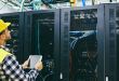 A man in a yellow hard hat and yellow vest checks cables in a server farm.
