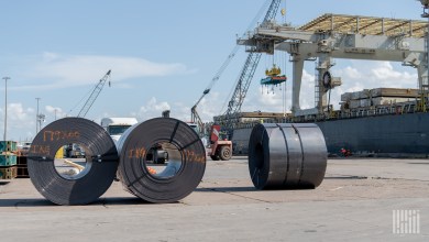 Steel coils sit on a port dock, with a truck, crane and cargo ship behind them, as factory backlogs rise and steel costs climb.