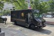 A brown UPS truck parked on a shaded city street, as seen from the side.