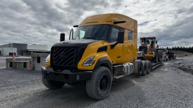Yellow Volvo VNX sleeper tractor hauling a Volvo wheel loader on a lowboy trailer across a gravel lot