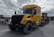 Yellow Volvo VNX sleeper tractor hauling a Volvo wheel loader on a lowboy trailer across a gravel lot