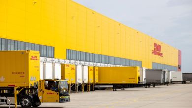 A large DHL warehouse with a yellow facade and yellow trucks at the dock doors.