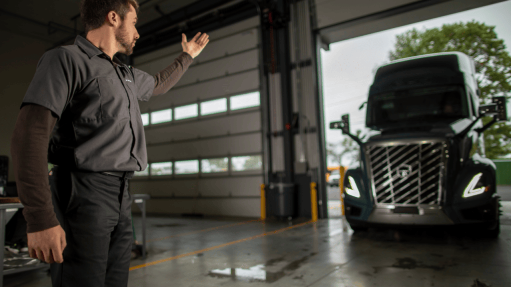 A man in a work uniform gestures toward a Volvo semitruck at the open door of a dealer service bay