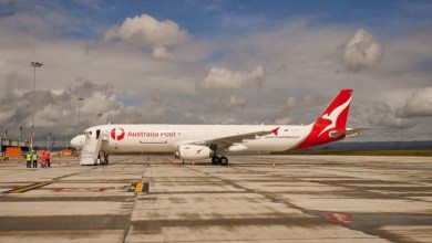 Side view of a red-tailed Australia Post-Qantas Freight cargo jet on an airport parking area, with puffy clouds in the background