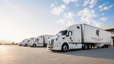 Four white Spartan Carrier Group tractor-trailers parked in a row outside a warehouse under a blue sky
