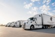Four white Spartan Carrier Group tractor-trailers parked in a row outside a warehouse under a blue sky